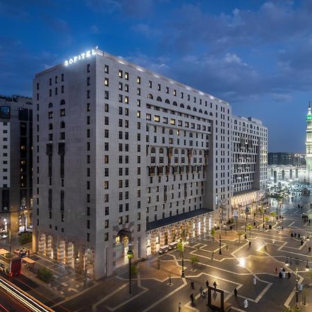 Sofitel Shahd Al Madinah Otel Medine Dış mekan fotoğraf The photo shows a large modern building, likely a hotel, situated in a cityscape at dusk. The structure has a distinct architectural style with multiple stories and illuminated windows. Surrounding the building is a plaza or open area with trees and 