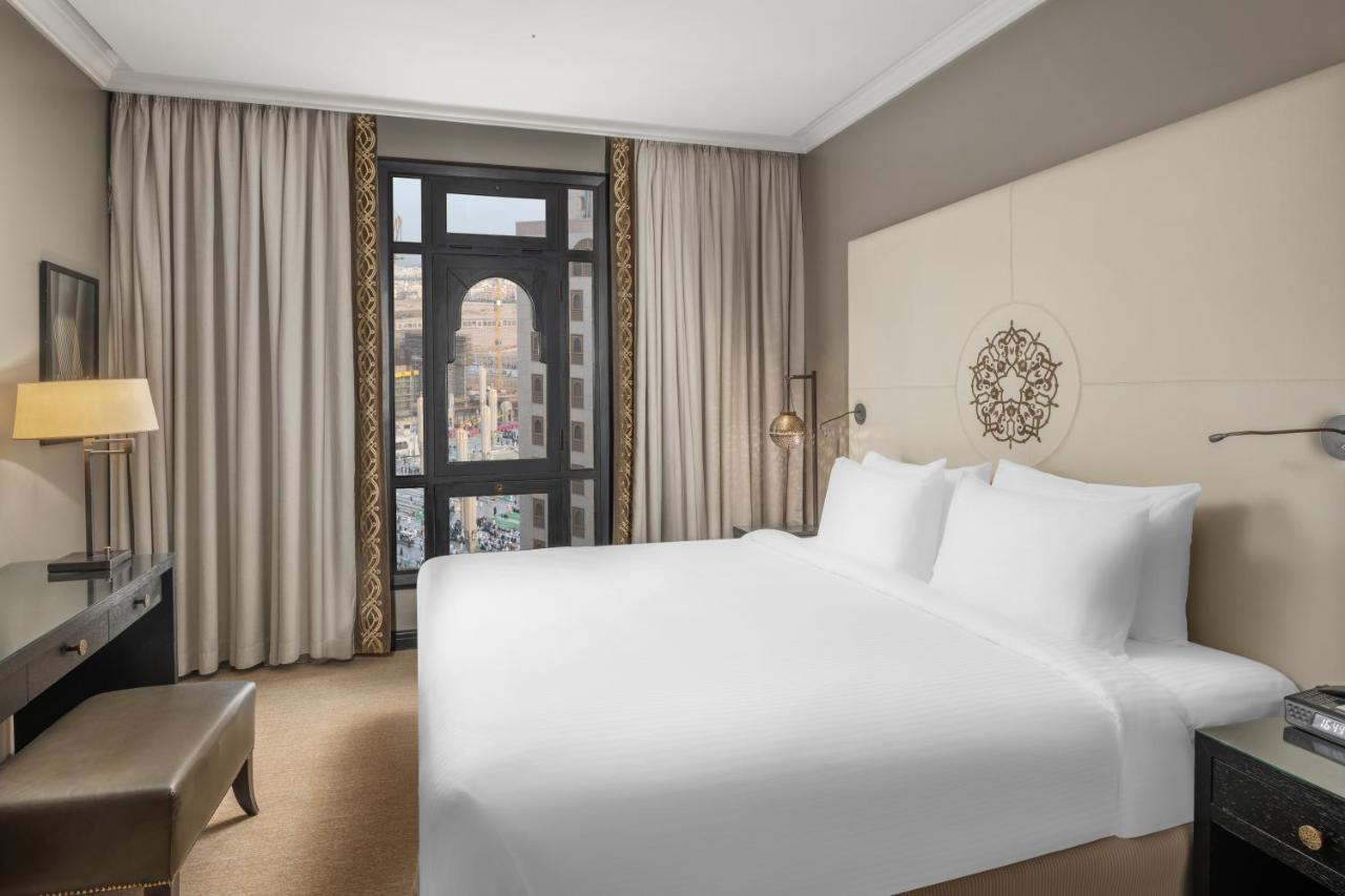 Sofitel Shahd Al Madinah Otel Medine Dış mekan fotoğraf The photo shows a modern hotel room featuring a large bed with white linens. There are beige walls and plush carpet, contributing to a clean and contemporary aesthetic. Large curtains frame a window that provides a view of the outside, likely a citys