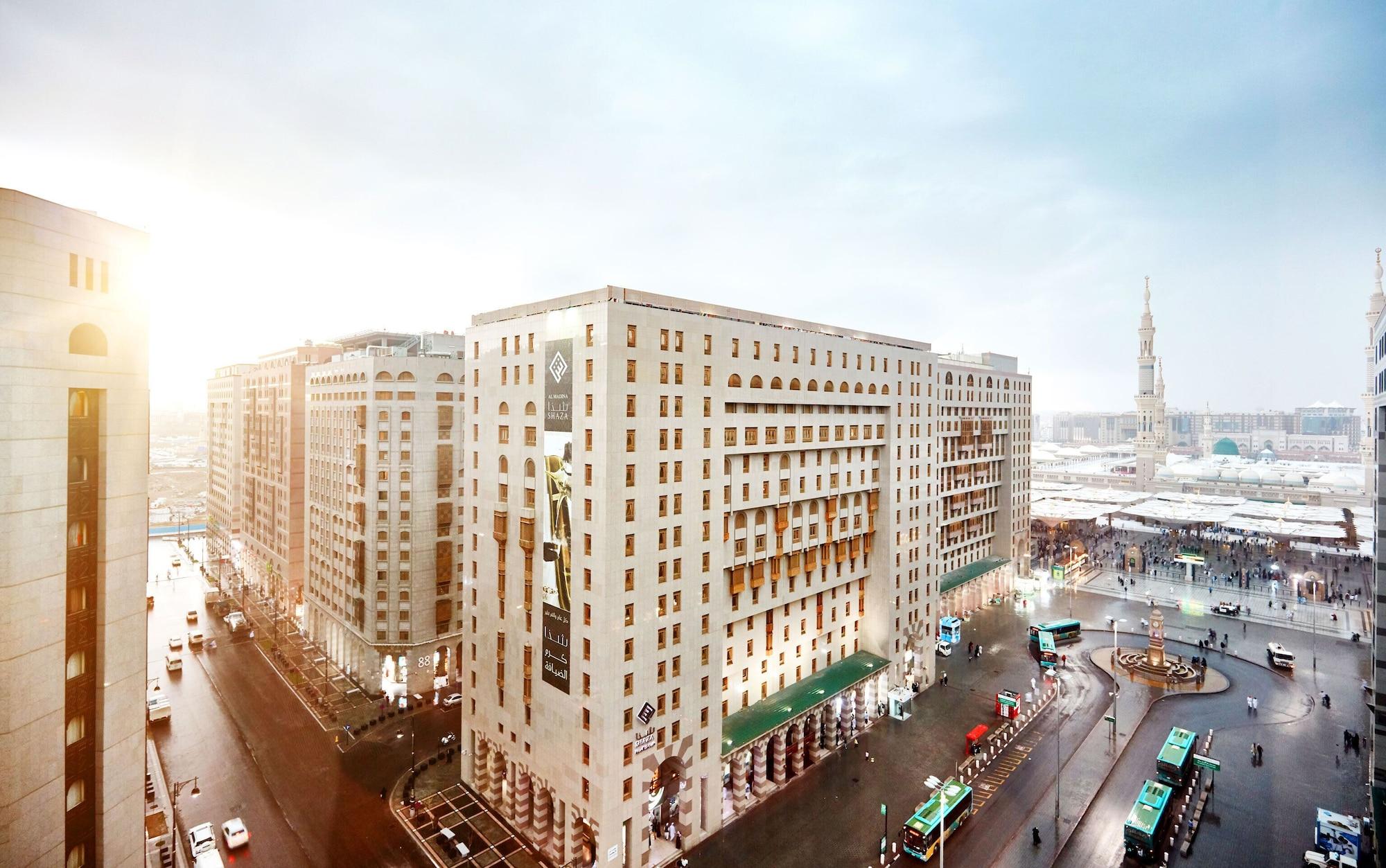 Sofitel Shahd Al Madinah Otel Medine Dış mekan fotoğraf The photo shows an urban landscape featuring several large buildings, which appear to have a modern architectural style. The scene is depicted during a time of day when sunlight is visible, suggesting either early morning or late afternoon. In the fo