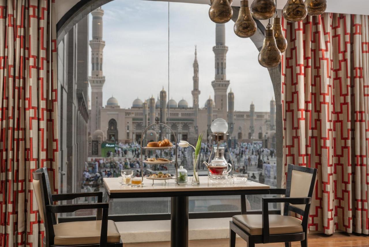 Sofitel Shahd Al Madinah Otel Medine Dış mekan fotoğraf The photo shows a dining setup inside a room with a large window. A table is set for a meal, featuring a tiered stand with various foods, a teapot, and cups. The room is decorated with elegant furniture and patterned curtains. Through the window, the