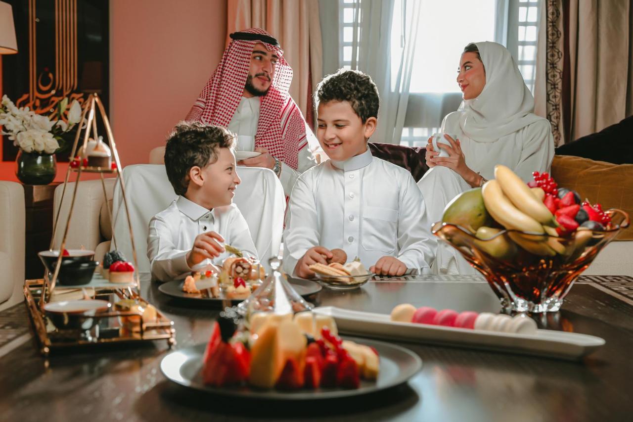 Sofitel Shahd Al Madinah Otel Medine Dış mekan fotoğraf The photo shows a warm family moment featuring two young boys, dressed in traditional attire, engaged in a joyful conversation. They are seated around a table adorned with various dishes, including an array of fruits and pastries. In the background, 