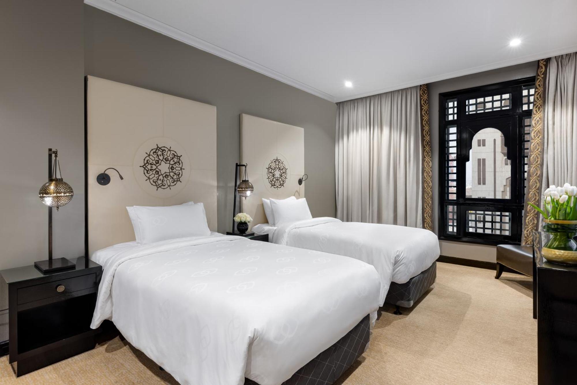 Sofitel Shahd Al Madinah Otel Medine Dış mekan fotoğraf The photo shows a modern hotel room featuring two twin beds with white linens. Each bed is flanked by bedside tables with stylish lamps. The walls are painted in a neutral tone, and there are decorative panels behind the beds, adding a touch of elega