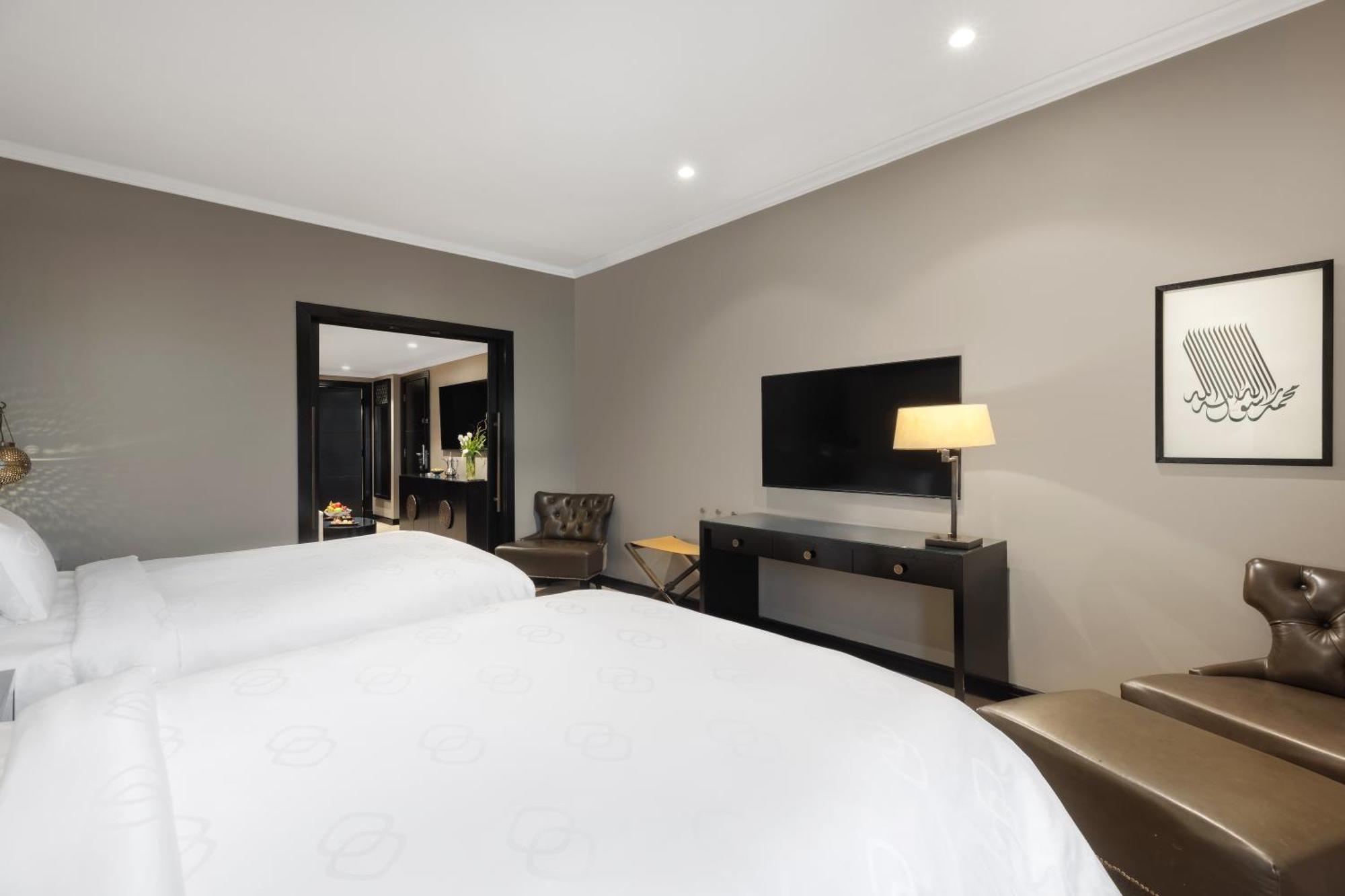 Sofitel Shahd Al Madinah Otel Medine Dış mekan fotoğraf The photo shows a modern hotel room with a neutral color palette. It features two beds with white linens, a stylish lamp on a side table, and a flat-screen TV mounted on the wall. There is a dark wood console table beneath the TV, and a comfortable a