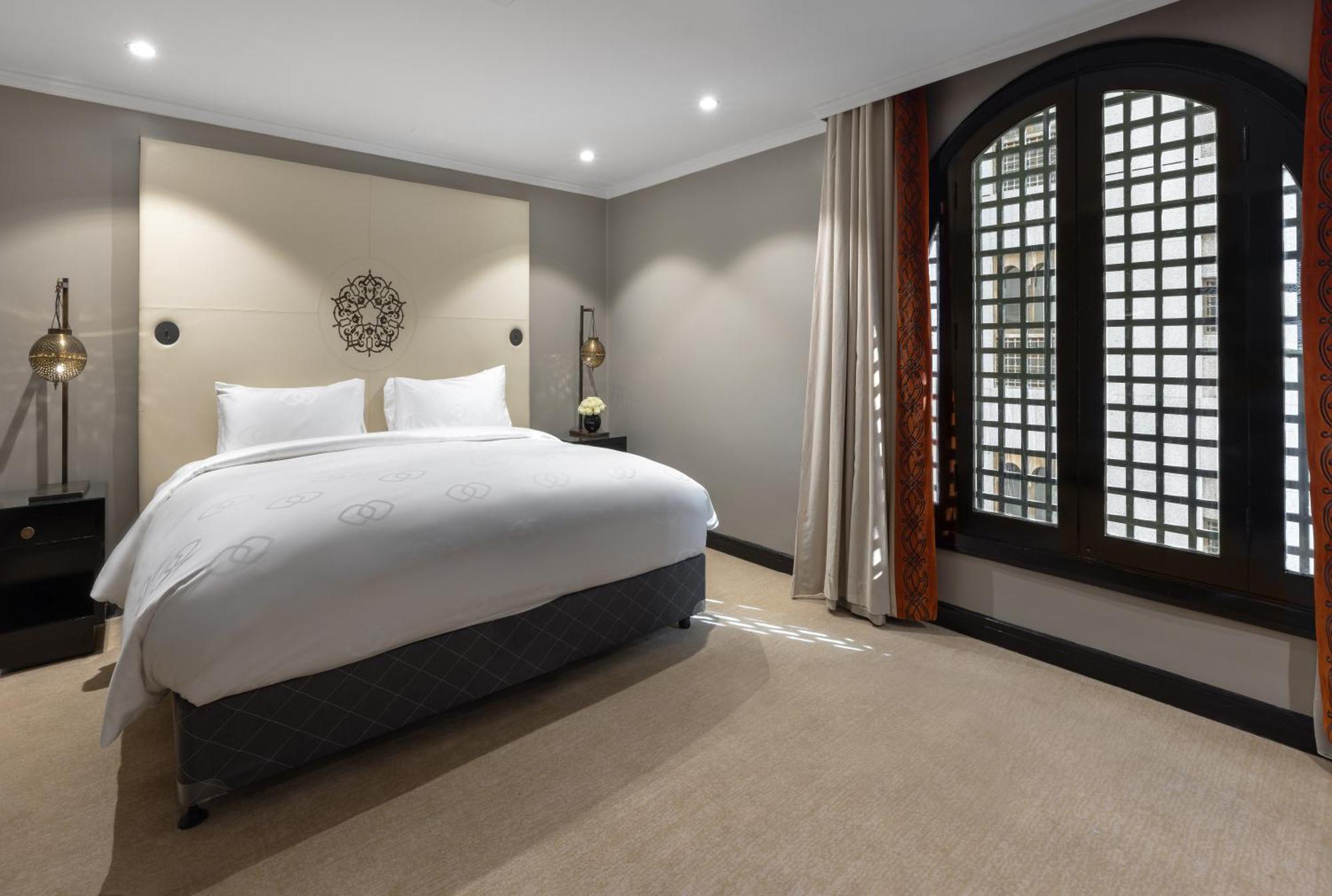 Sofitel Shahd Al Madinah Otel Medine Dış mekan fotoğraf The photo shows a modern hotel bedroom. It features a large bed with white bedding and two pillows. The walls are painted in neutral tones, and there is decorative wall art above the bed. To the right, there are large windows with patterned shutters,