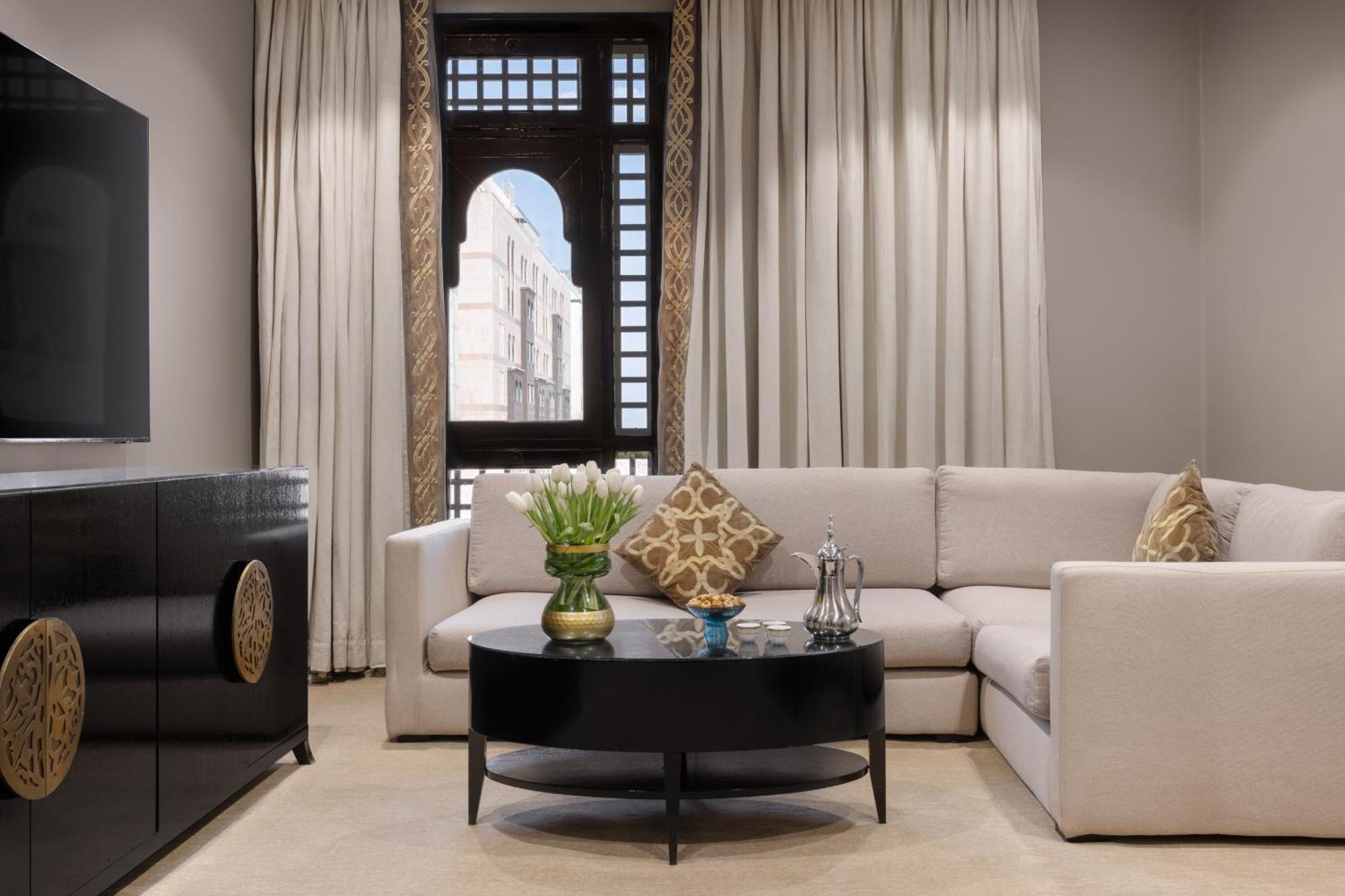 Sofitel Shahd Al Madinah Otel Medine Dış mekan fotoğraf The photo shows a stylish and modern living room. It features a light-colored sectional sofa with decorative cushions, positioned around a round black coffee table. On the table, there’s a vase with flowers and some decorative items. The room has lar