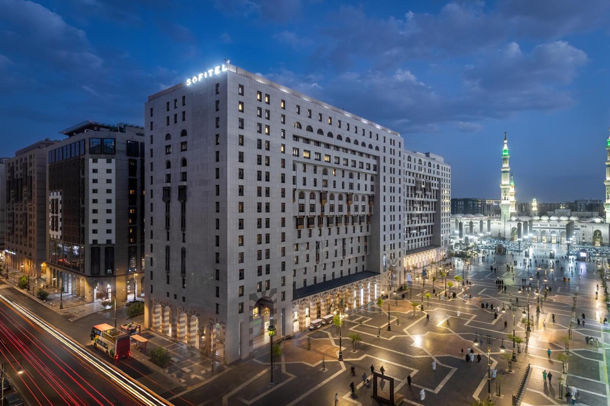 Sofitel Shahd Al Madinah Otel Medine Dış mekan fotoğraf The photo shows a large modern building, likely a hotel, situated in a cityscape at dusk. The structure has a distinct architectural style with multiple stories and illuminated windows. Surrounding the building is a plaza or open area with trees and 