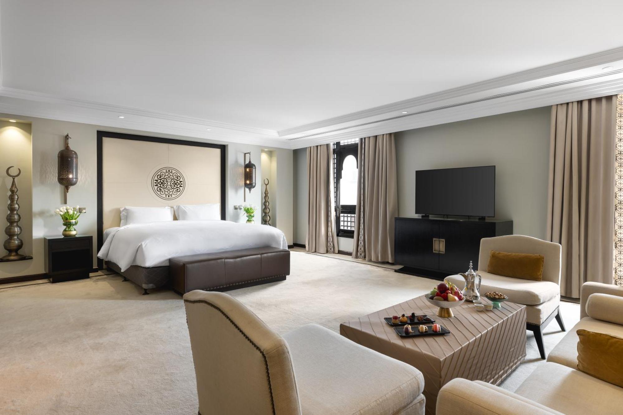 Sofitel Shahd Al Madinah Otel Medine Dış mekan fotoğraf The photo shows a spacious and stylish hotel room. The room features a large bed with a dark headboard, complemented by light-colored bedding. There are two comfortable armchairs facing a coffee table, which holds some snacks and a drink. A modern te