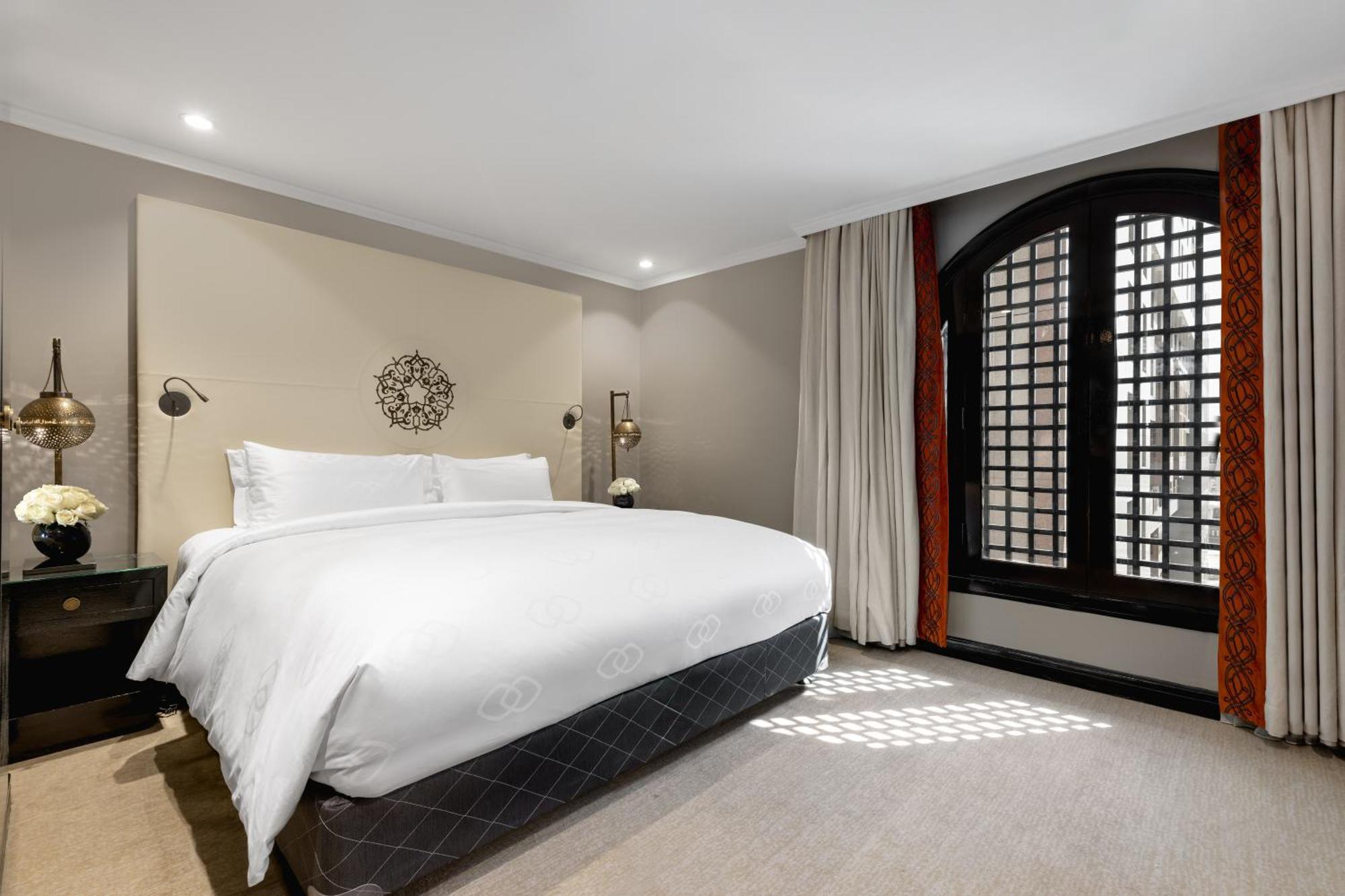 Sofitel Shahd Al Madinah Otel Medine Dış mekan fotoğraf The photo shows a stylish hotel room featuring a large bed with white bedding positioned against a light-colored wall. Decorative elements include a circular wall piece above the bed and bedside lamps. The room has large windows with a unique grid pa